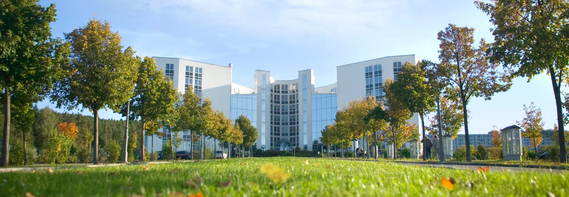 Modernes, mehrstöckiges Gesundheitszentrum der Eckert Schulen, umgeben von einer Allee mit Bäumen und gepflegten Rasenflächen, unter blauem Himmel.