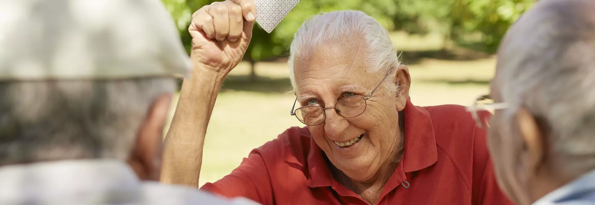 Ein älterer Mann in rotem Polohemd zeigt lachend eine Spielkarte in die Höhe, während er mit zwei weiteren Senioren draußen Karten spielt.